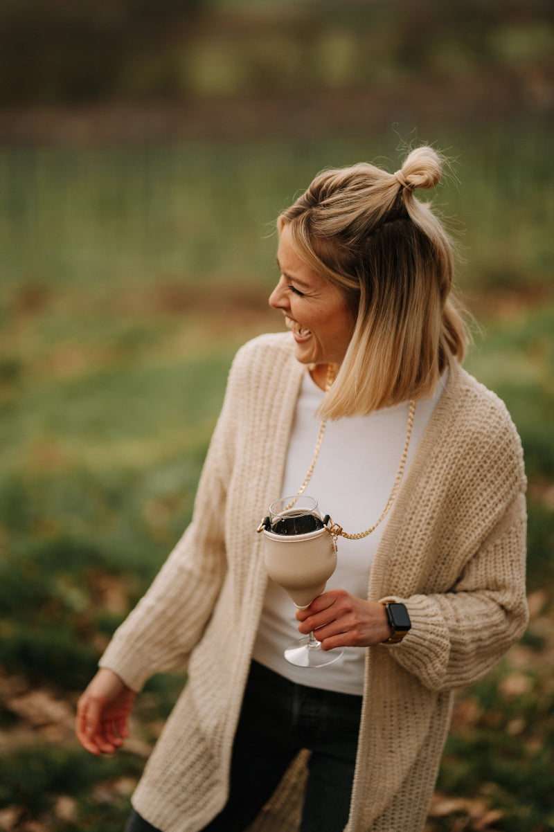 Eine Person steht im Freien und hält ein Getränk in einem Glas. Sie trägt eine beige Strickjacke, ein weißes Oberteil und eine schwarze Hose. Im Hintergrund ist eine grüne Landschaft erkennbar.
